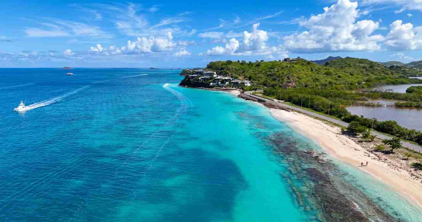Aerial view of a tropical beach with turquoise water, white sand, and green hills with houses along the shoreline.