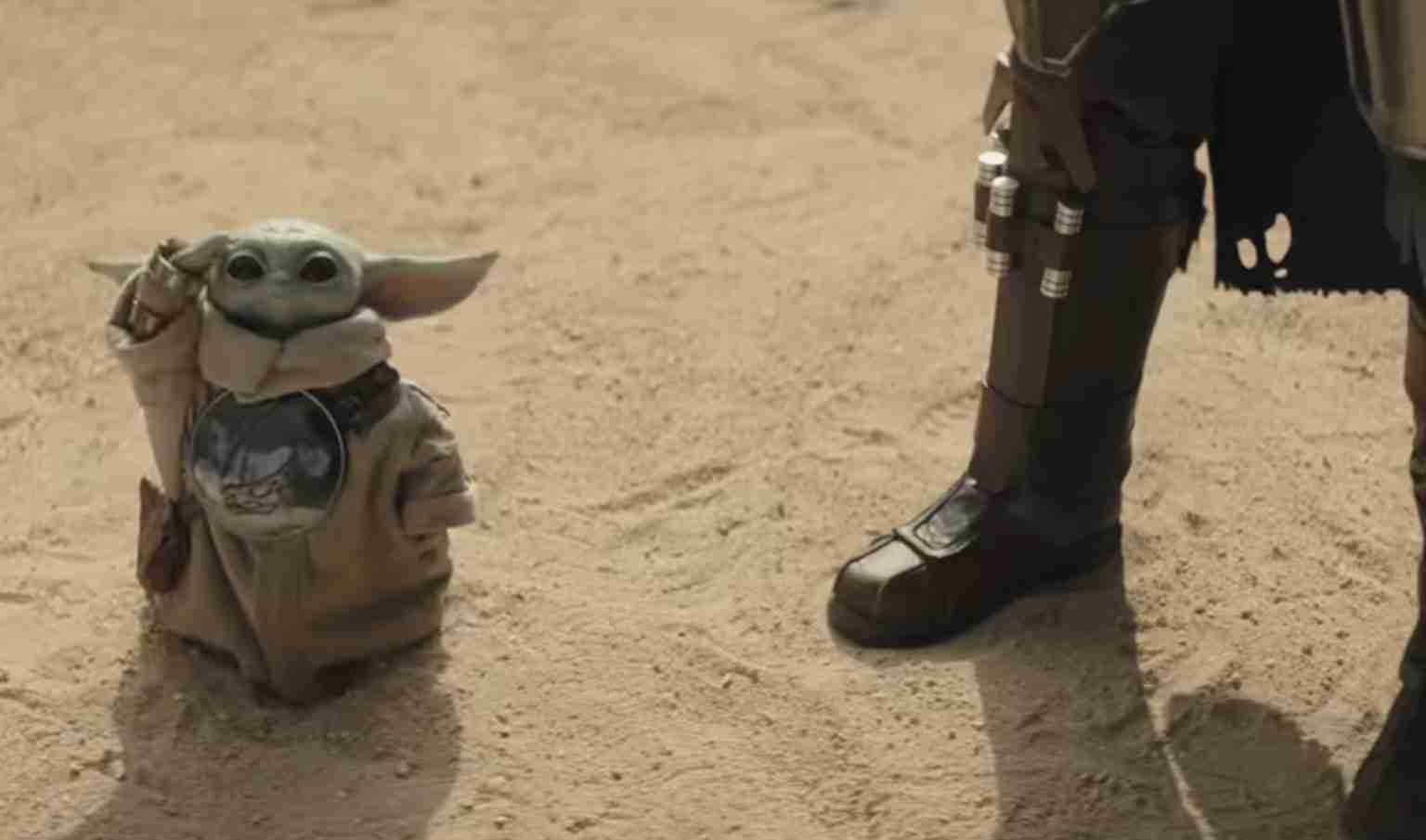 Grogu (Baby Yoda) in a beige robe stands in a desert beside a partial view of an armored person.