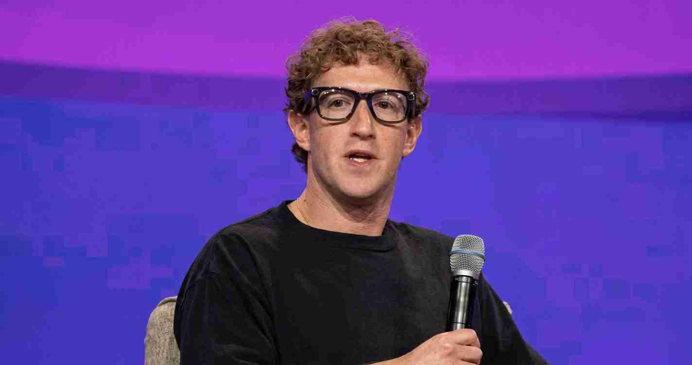 Man wearing a black shirt and glasses speaks into a handheld microphone on a purple stage backdrop.