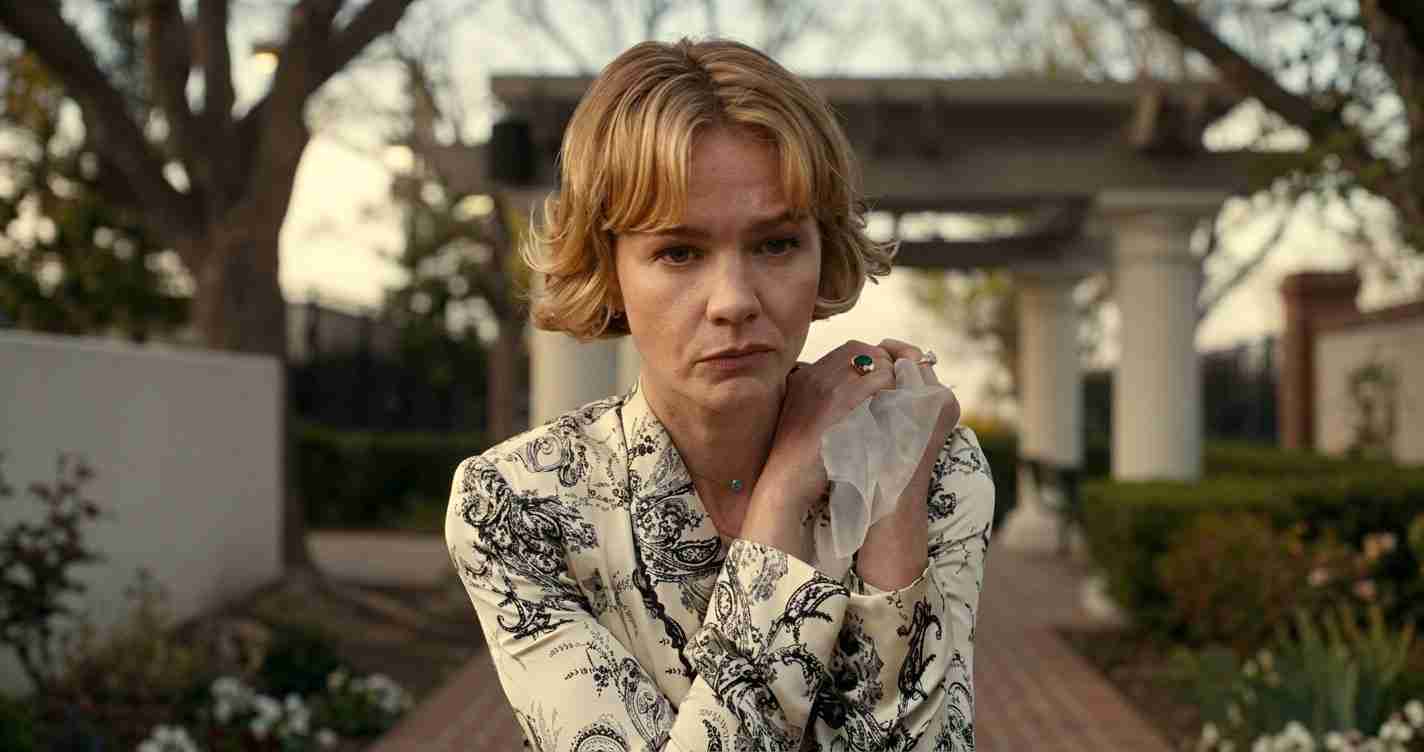 Woman with short blond hair in a patterned blouse, looking worried as she holds a napkin to her shoulder outdoors in a garden.