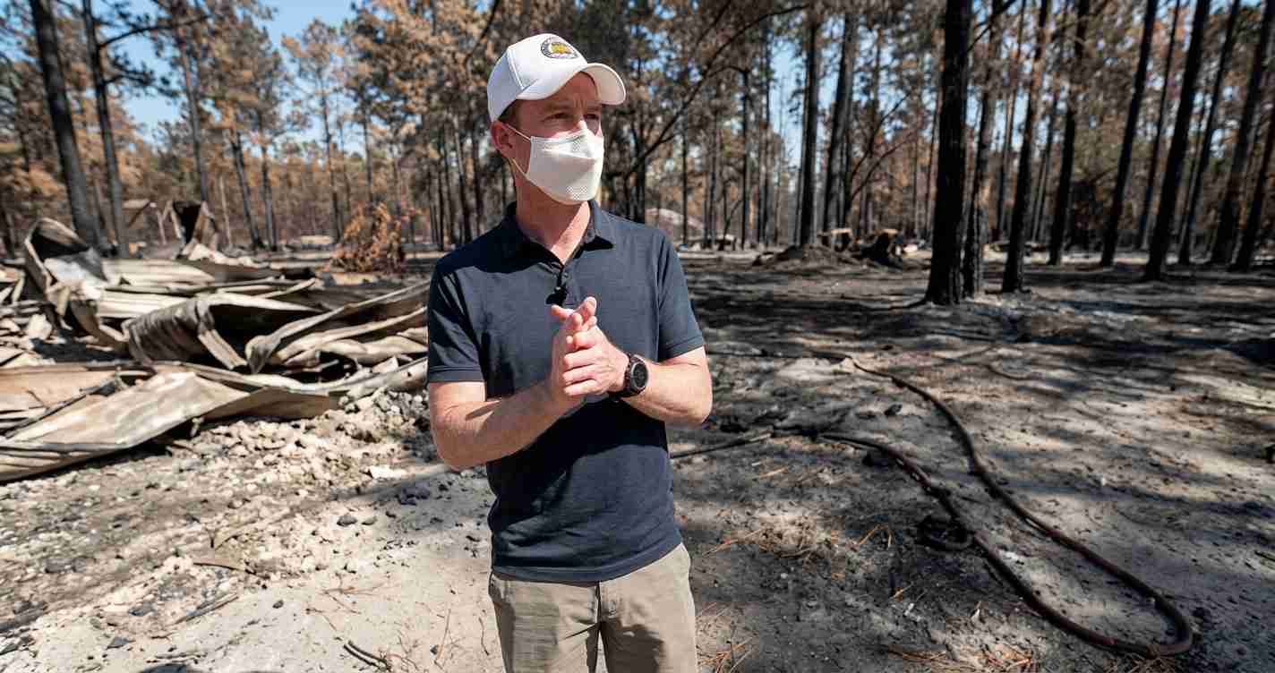 Man in a white cap and mask stands in a burned forest, hands clasped, amid charred debris and scorched trees.