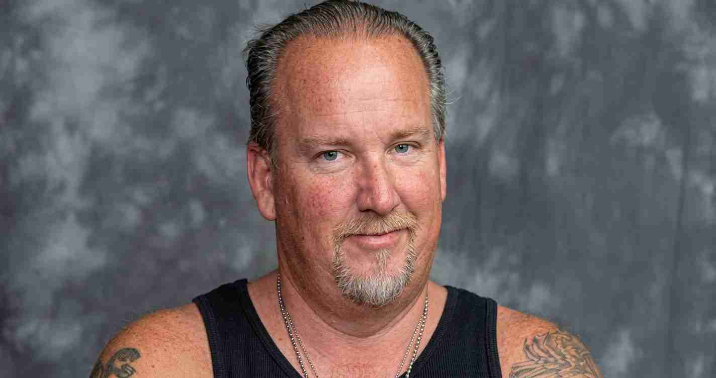 Head-and-shoulders portrait of a man with short gray hair, facial stubble, wearing a black tank top, against a gray mottled backdrop
