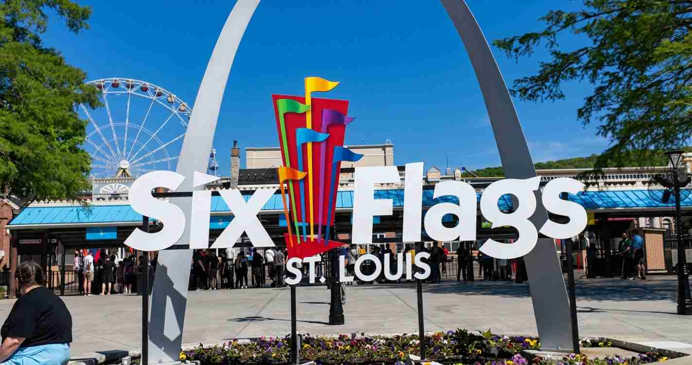 Six Flags St. Louis entrance sign with a colorful sculpture and a Ferris wheel in the background under a blue sky, with visitors nearby.