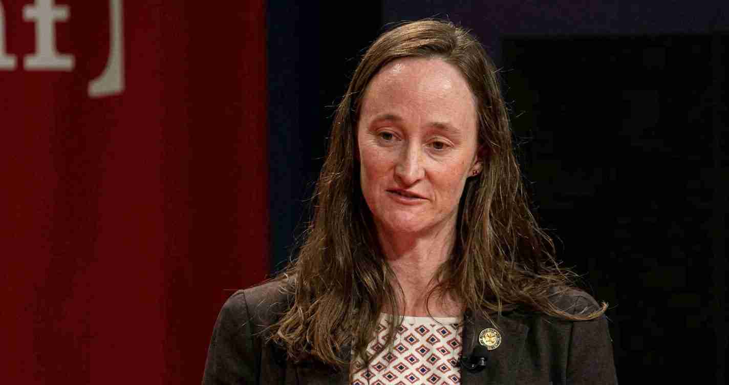 Woman in a dark blazer speaking on a stage, wearing a patterned blouse and a gold lapel pin, against a red and dark backdrop.