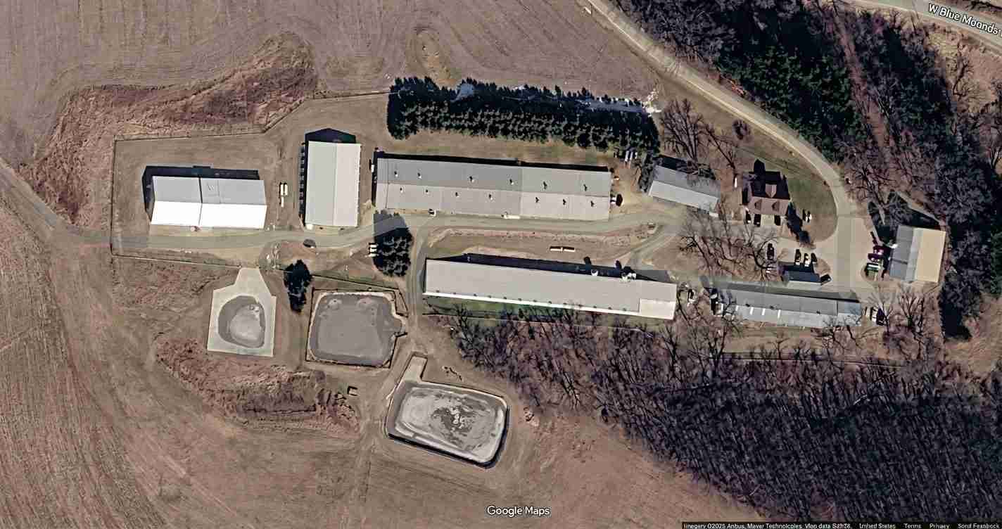 Aerial view of a rural industrial site with several long white-roofed buildings, roads, and clusters of trees surrounding multiple rectangular basins or ponds.