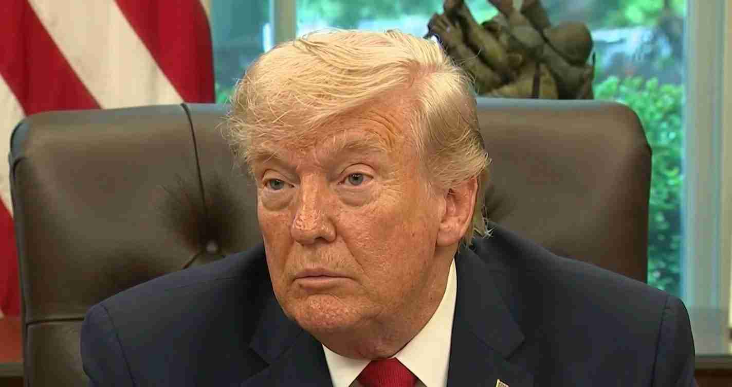 Close-up of an older man in a dark suit and red tie seated in an office chair with an American flag in the background.