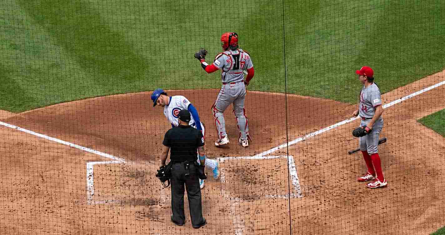 Baseball scene at home plate: an umpire in black stands near the catcher (blue helmet) and a baserunner in gray with red accents near the plate as a game unfolds.