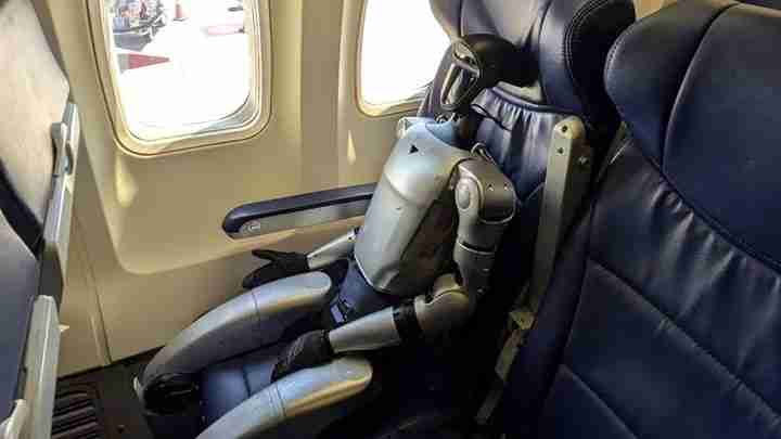 Silver humanoid robot seated in an airplane window seat, strapped in with arms resting in its lap.