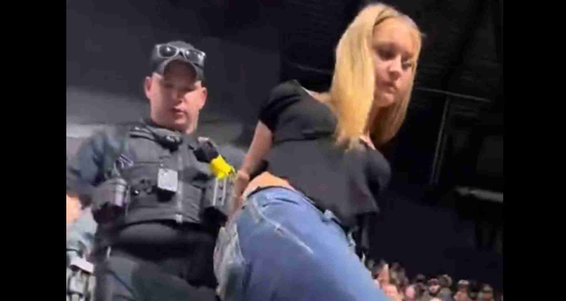 Security guard in tactical vest and cap standing on stage beside a blonde woman leaning toward the audience below.