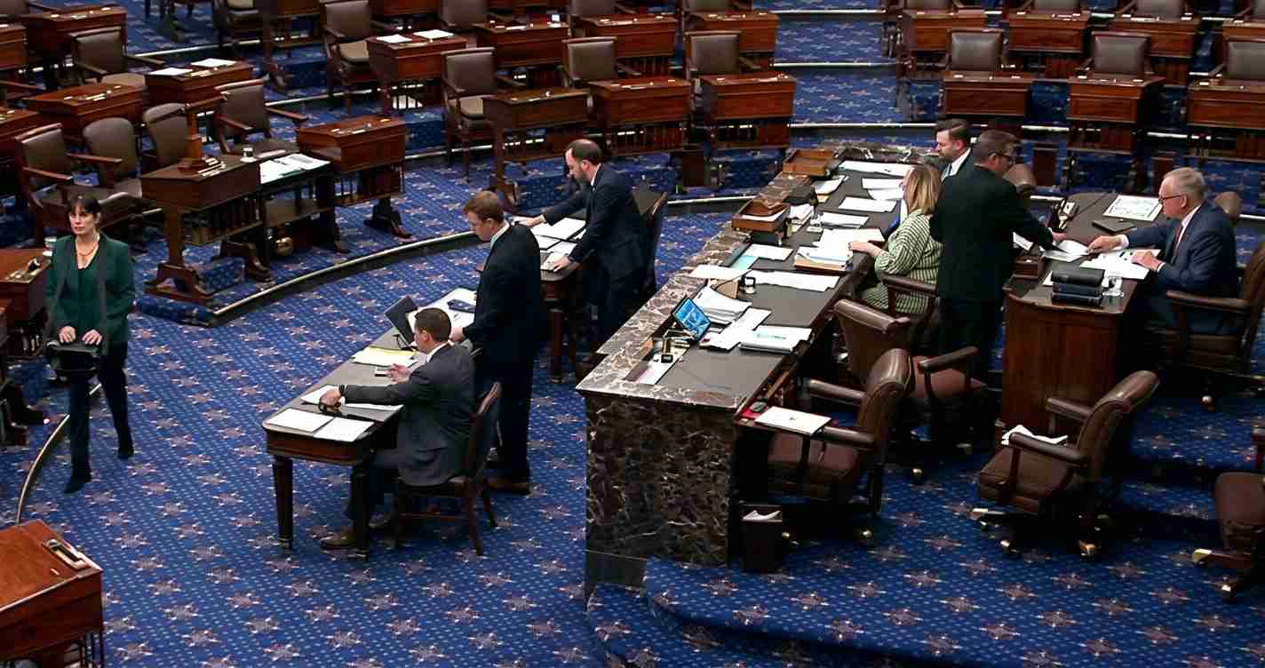 Legislative chamber with lawmakers at desks around a central marble podium on a blue carpeted floor.