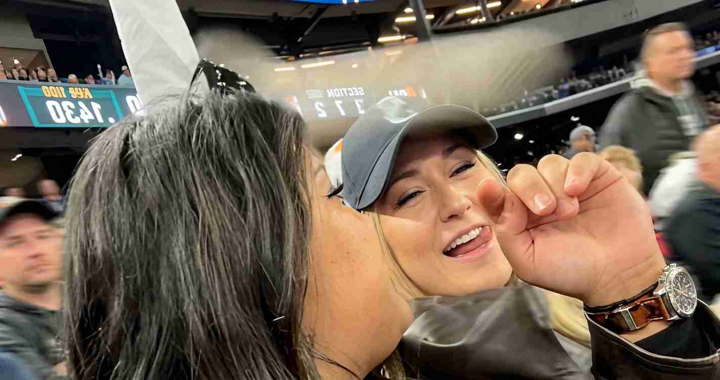 Two people share a playful kiss at a crowded stadium, with a gray baseball cap-wearing friend smiling and a metal-band watch visible on the foreground wrist.