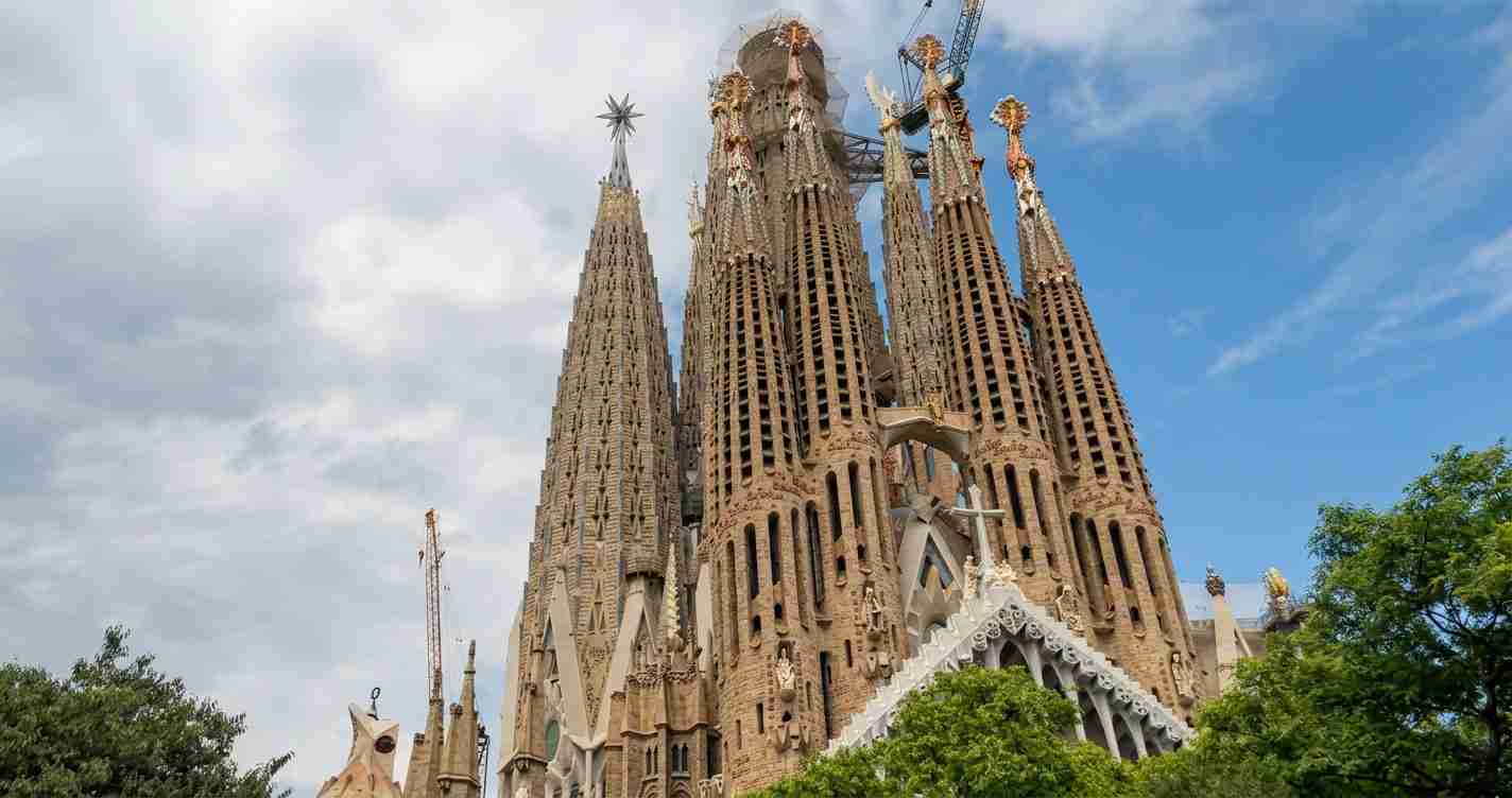 Sagrada Família basilica in Barcelona with tall stone spires reaching into a blue sky, surrounded by green trees