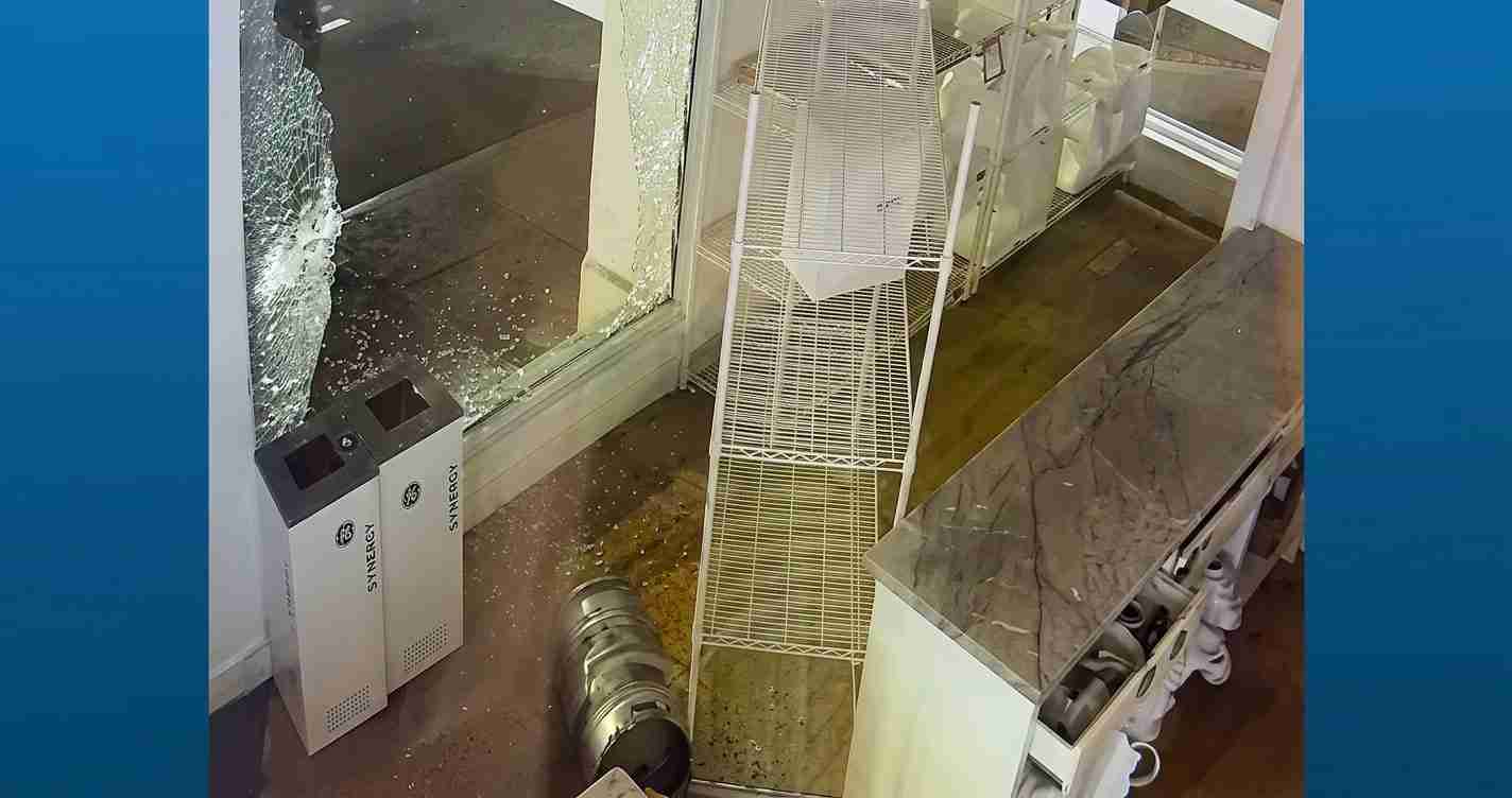 Store interior after an incident: broken storefront window, shattered glass, and fallen white wire racks with debris on the floor.