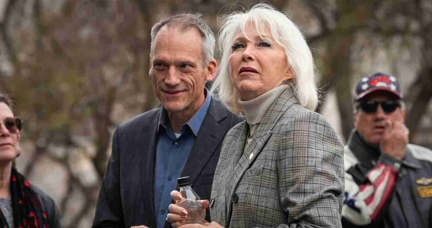 Two older adults outdoors: a woman with white hair in a gray plaid blazer holding a water bottle, beside a man in a blue shirt and dark jacket.