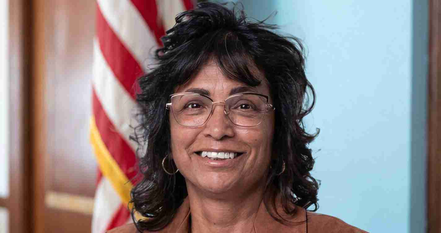 Smiling woman with short dark hair and glasses, standing in front of an American flag.