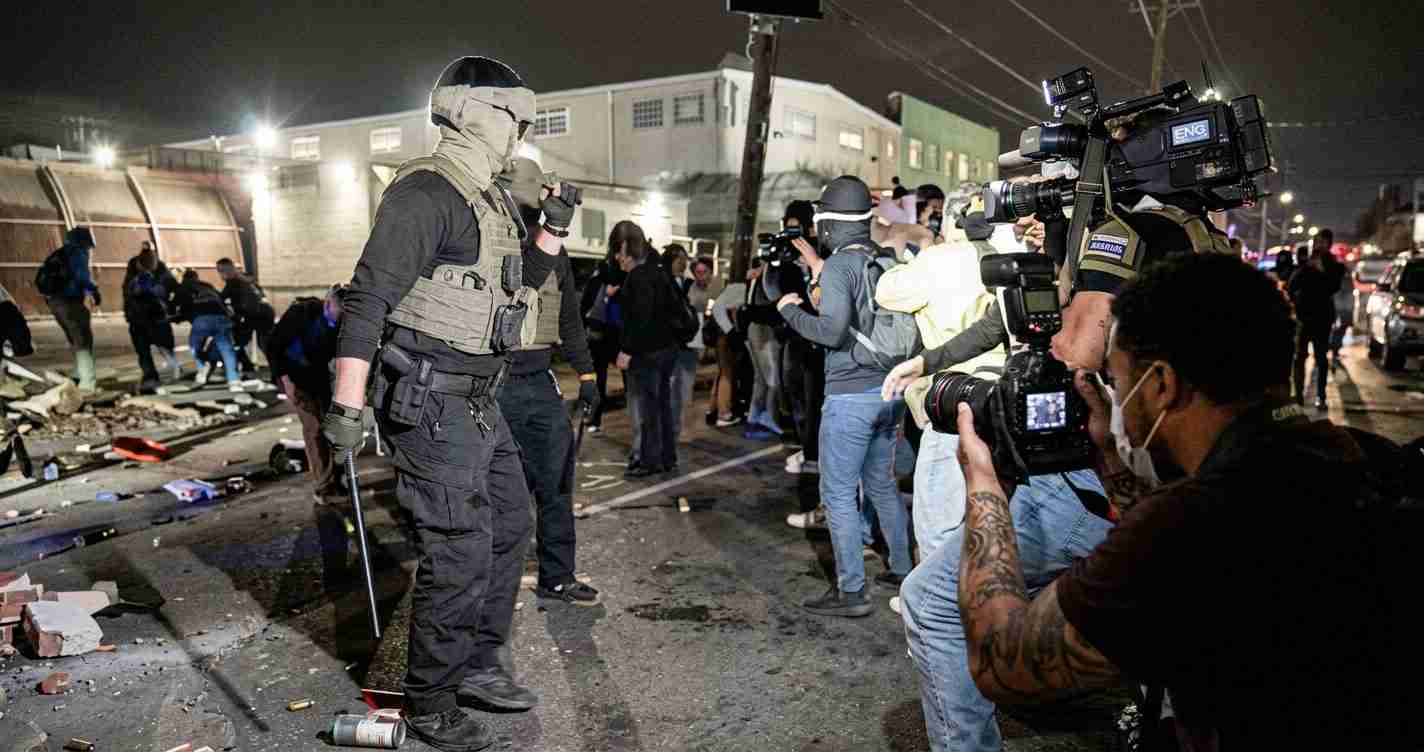Armored officer in tactical vest and helmet stands on a debris-strewn street at night as photographers document the scene.