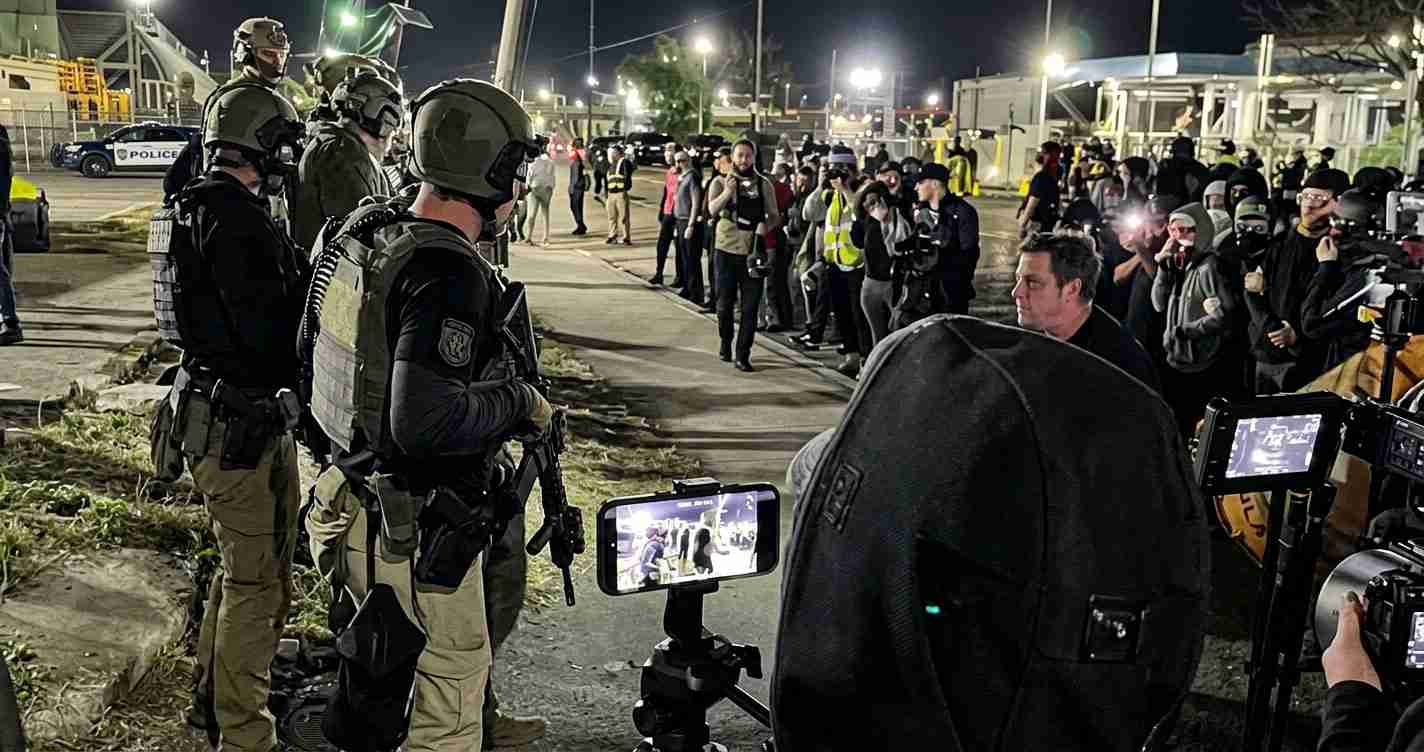 Line of heavily armed police in tactical gear facing a crowd of journalists and bystanders at night, with cameras filming the scene and police cars in the background.