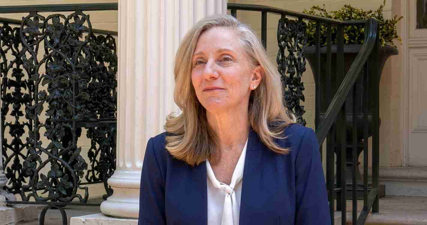 Portrait of a middle‑aged woman with blonde hair, wearing a navy blazer and white blouse, seated outside a columned building with ornate iron railing.