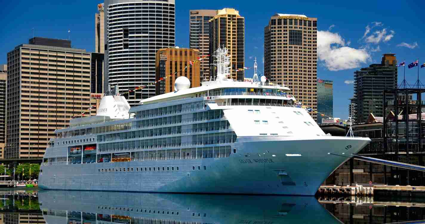 Large white cruise ship docked at a city harbor with skyscrapers in the background, on a sunny day.