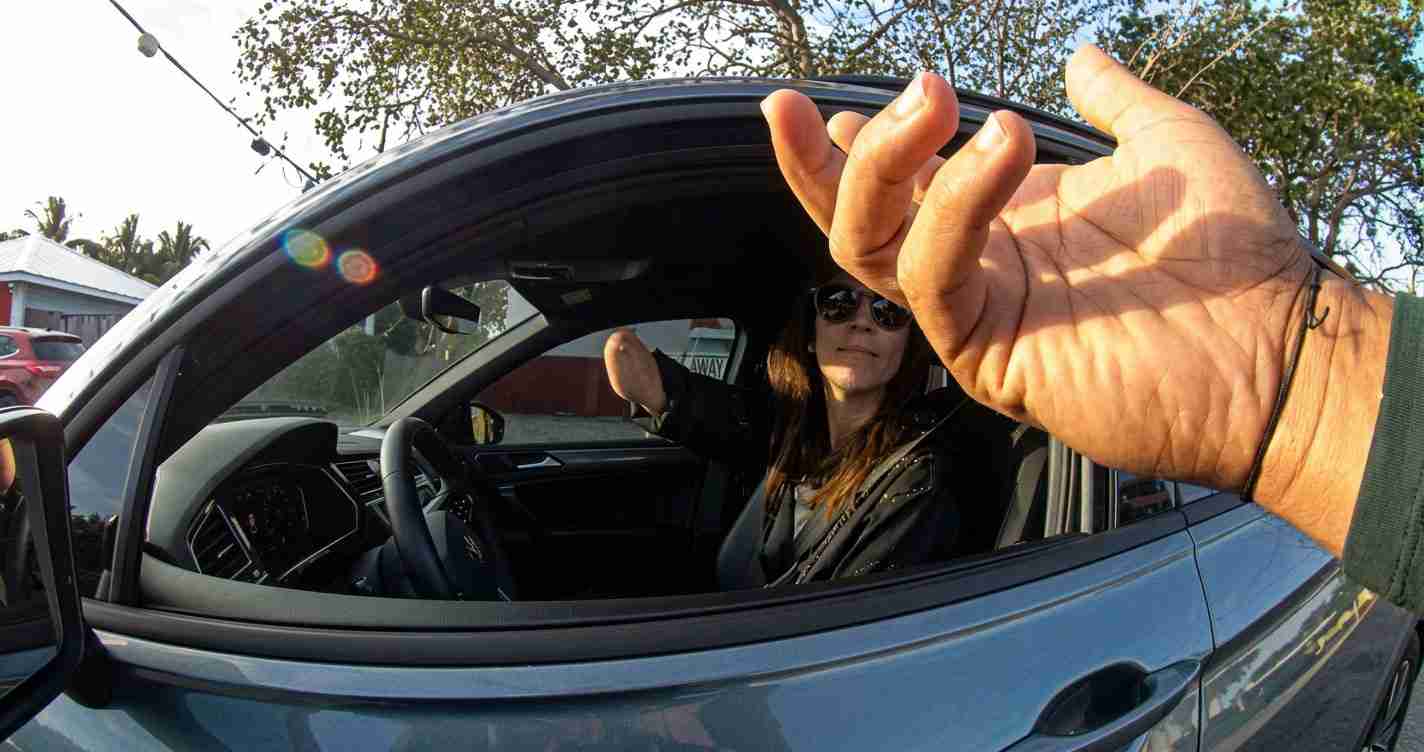 Driver in a blue car with the window down, wearing sunglasses, looks at the camera; a large hand is in the foreground reaching toward the window.