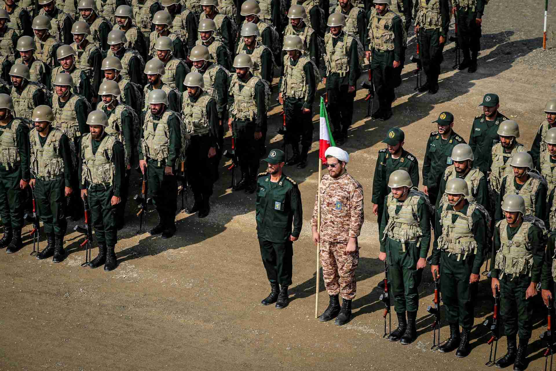 Line of soldiers in olive uniforms and helmets standing at attention, two front-line officers with an Iranian flag nearby