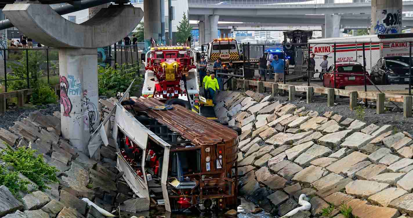 Bus wrecked and tipped onto its side in a rocky drainage trench, with emergency responders, rescue trucks, and bystanders along a fenced roadway.