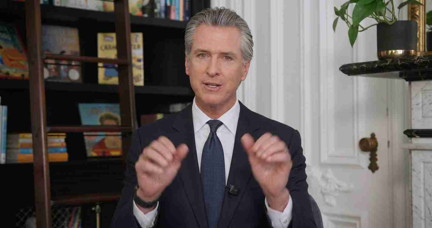 Man in a dark suit and tie speaks to the camera, hands gesturing mid-gesture in a library-studio backdrop.