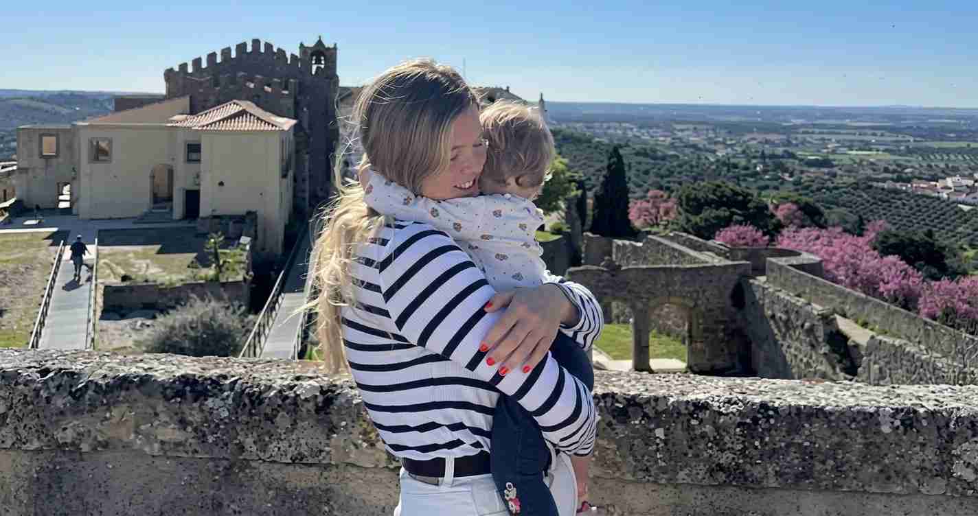 Woman in a striped top hugs a child on a stone railing at a hilltop fortress, with a valley and pink flowering trees in the background, on a sunny day.