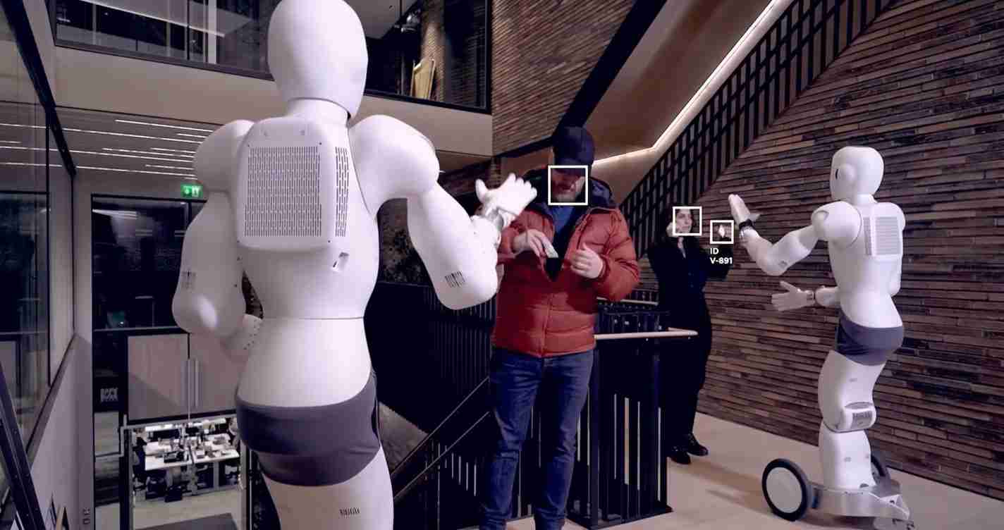 Two white humanoid robots gesture toward a man in an orange jacket on a balcony in a modern building interior.
