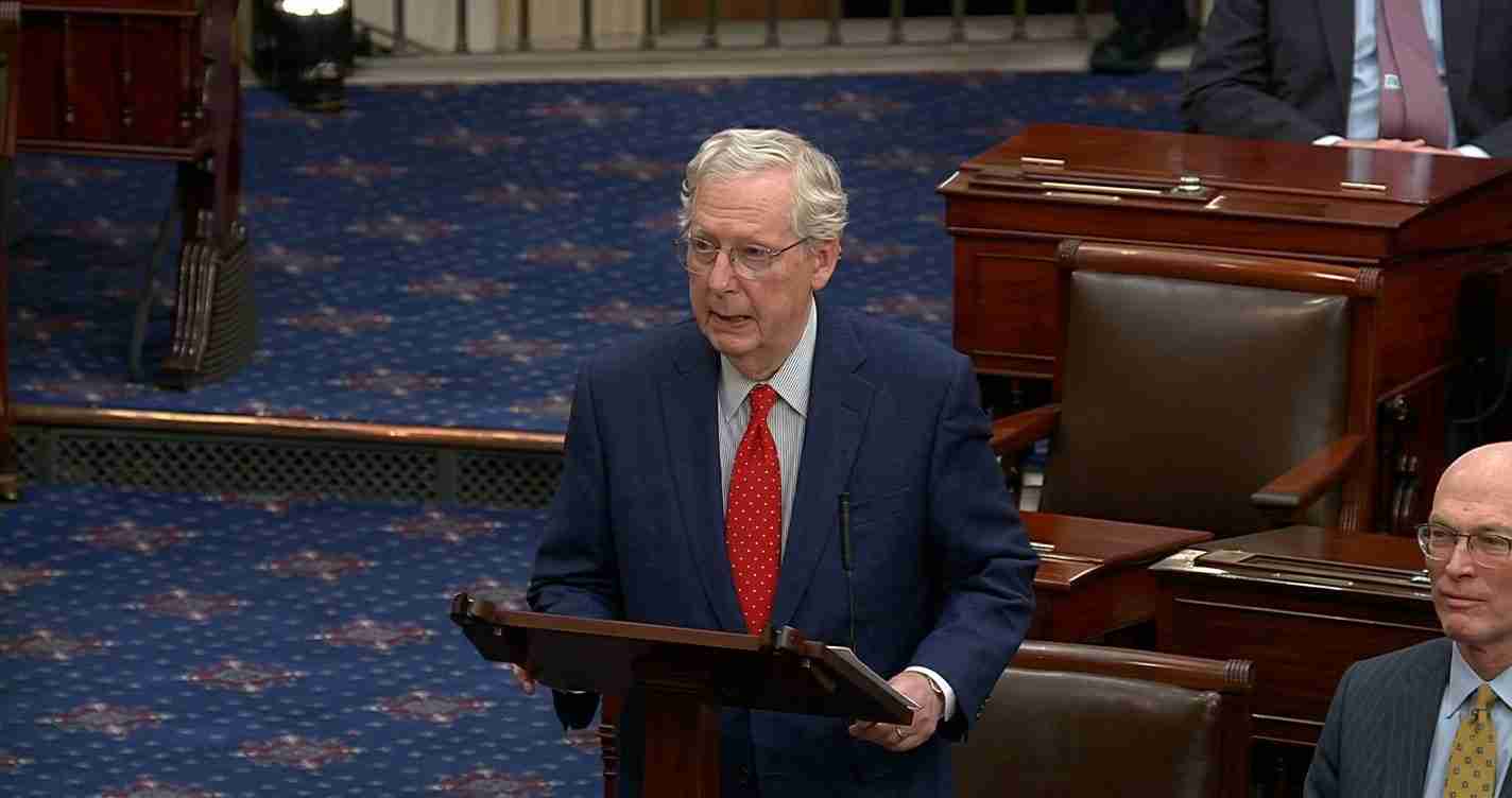 Older man in a navy suit and red polka-dot tie speaks from a wooden lectern in a formal chamber with blue carpeted floor.