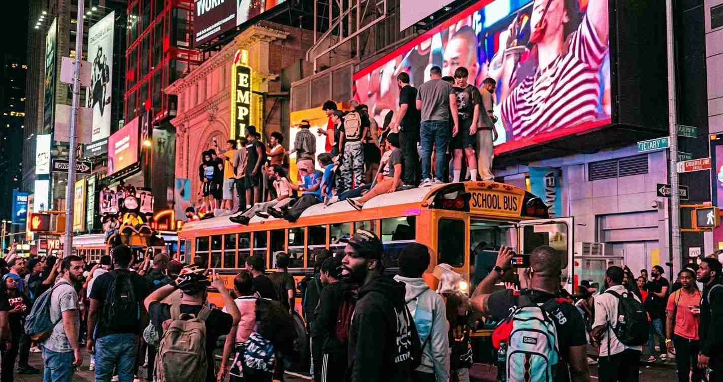Crowded city street at night with an orange school bus and many people sitting on its roof amid bright billboards.