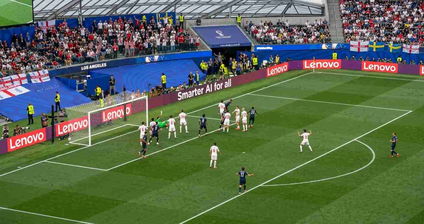 Soccer scene near the goal where white-team defenders contest a ball against attackers in dark uniforms during a set piece, with fans in the stands behind. Allied advertising boards line the pitch with Lenovo branding.