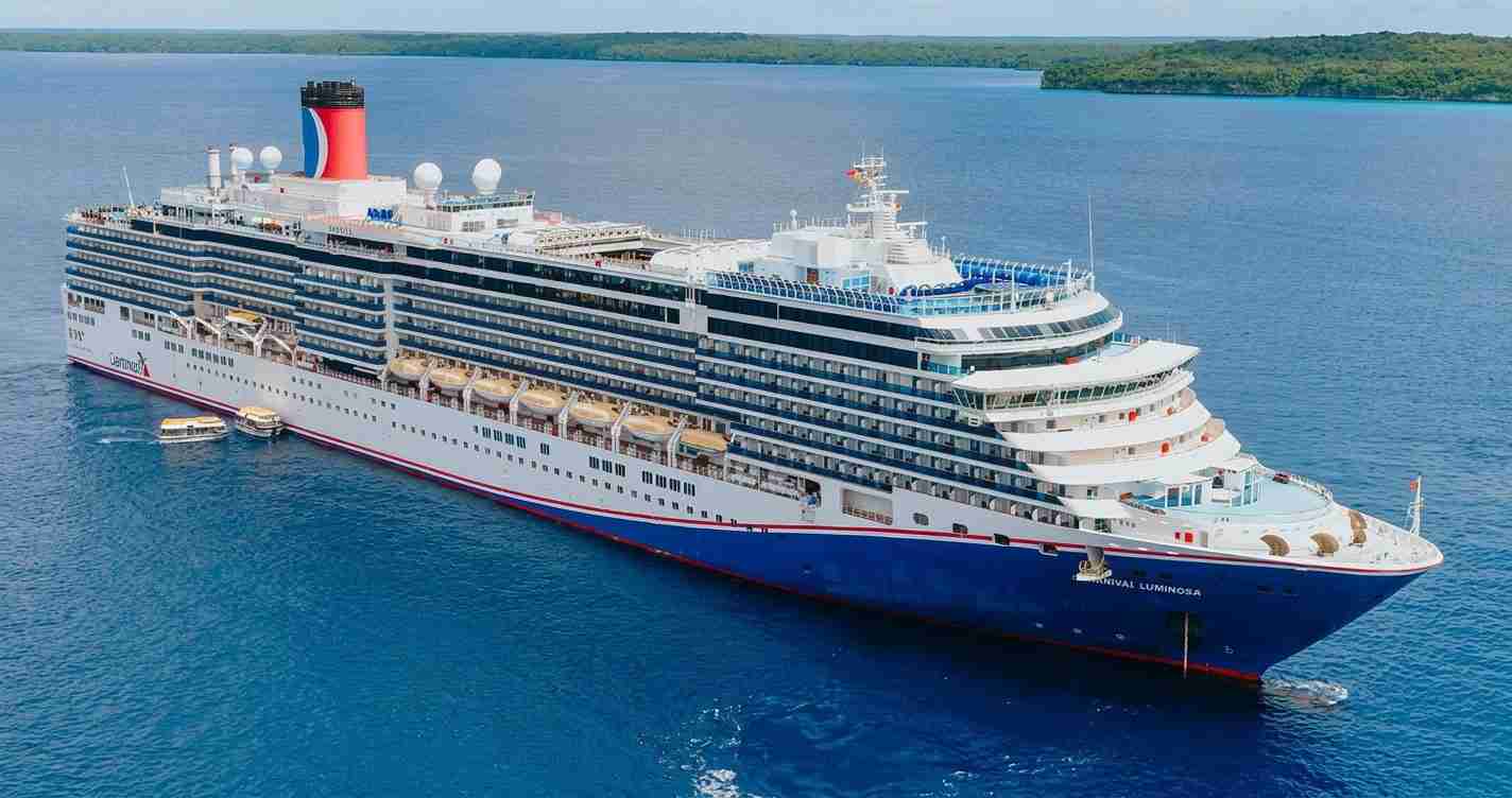 Large white-and-blue cruise ship on calm blue sea, viewed from above with multiple decks and lounge areas visible on the sides