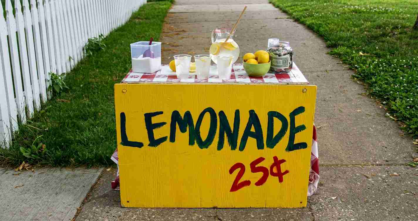 Lemonade stand on a sidewalk with a bright yellow sign reading LEMONADE 25¢ and a pitcher, cups, lemons, and a jar of money on top.