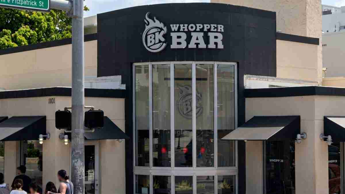 Exterior of Burger King Whopper Bar with large glass corner entrance and bold BK Whopper Bar sign on a dark facade, pedestrians nearby on a sunny day.