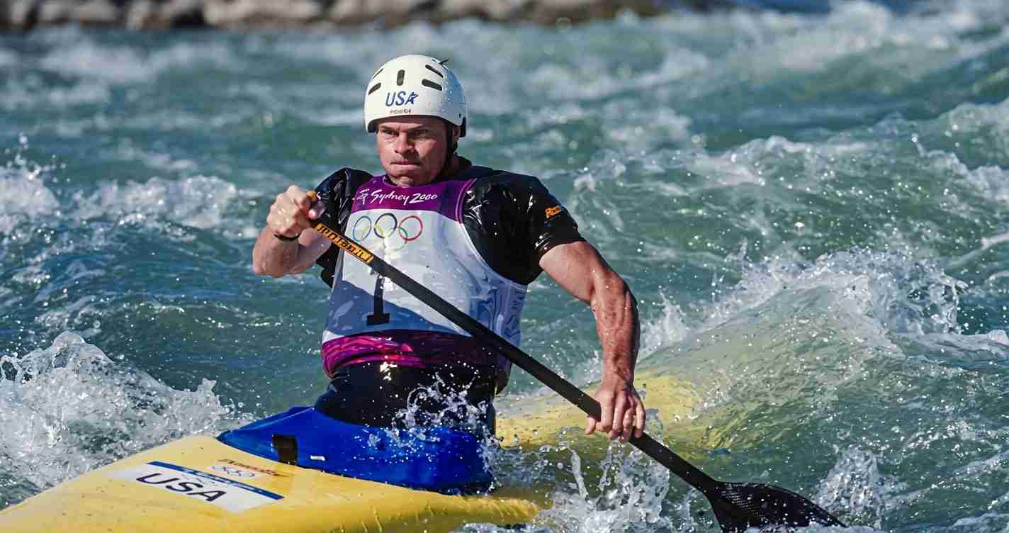 Male kayaker wearing a white helmet and USA vest paddling through rough whitewater in a yellow kayak with a black paddle.