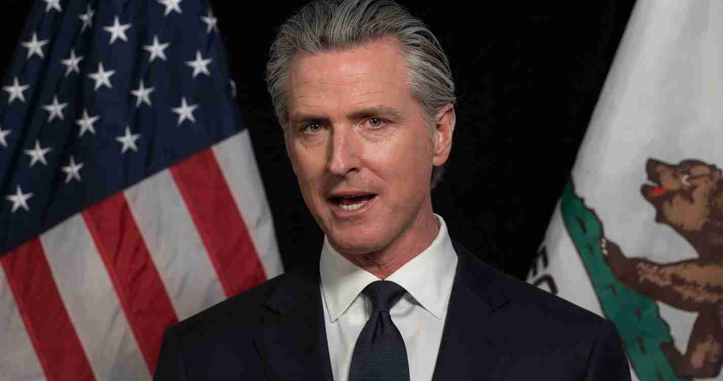 Man in a dark suit and tie speaks at a formal event, with American and state flags in the background.