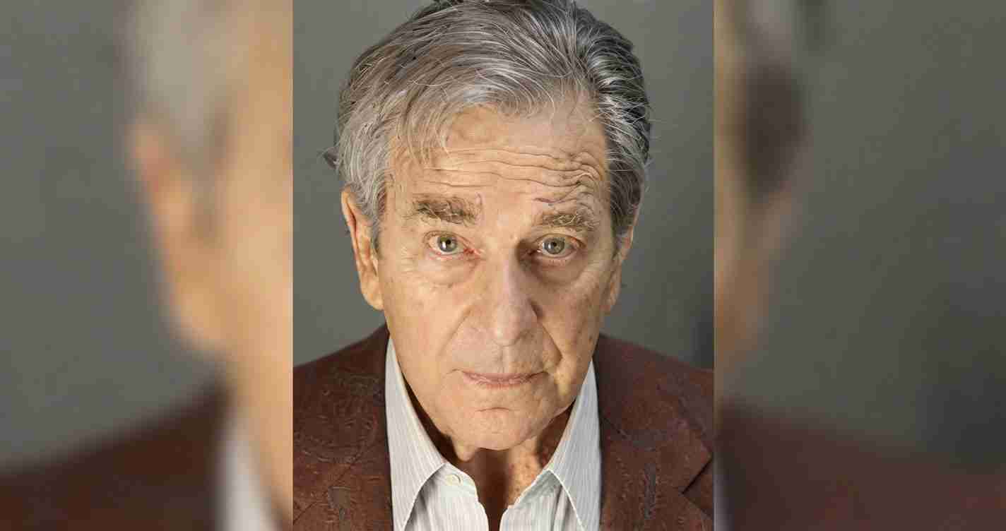 Older man with gray hair and a serious expression, wearing a brown blazer and light shirt, looking straight at the camera.