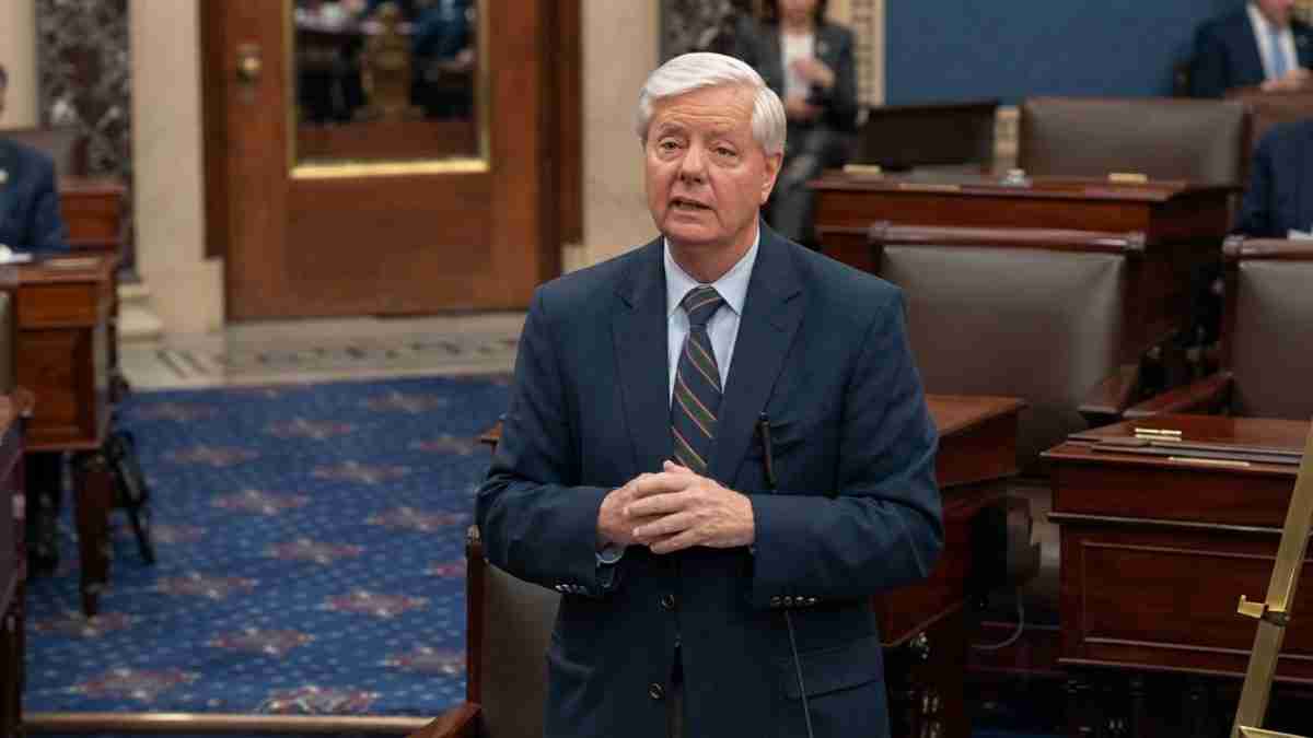 Older man in a navy suit and striped tie speaks in a formal legislative chamber with wooden desks and a blue carpet, hands clasped together.