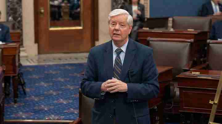Older man in a navy suit and striped tie speaks in a formal legislative chamber with wooden desks and a blue carpet, hands clasped together.