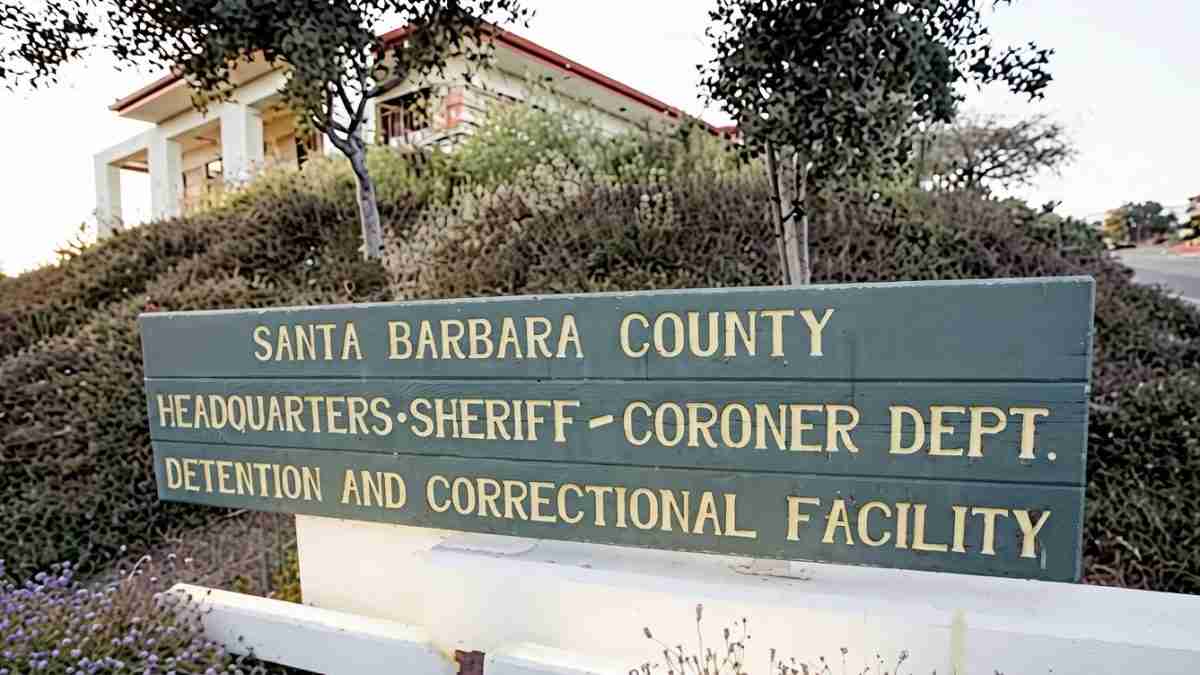 Green sign reading Santa Barbara County Headquarters Sheriff-Coroner Dept. Detention and Correctional Facility, with shrubbery and a building in the background.