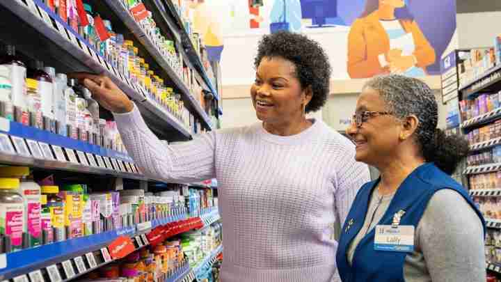 Shopper reaches for a product on a store shelf while a smiling associate watches nearby in a pharmacy aisle.